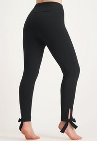 Svarta leggings i mjukt material med hög midja, figurnära design och ankelslitsar som accentueras av rosetter. Slät textur hela vägen.