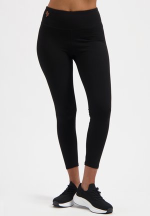Leggings noirs taille haute avec une texture lisse, une coupe ajustée, et un petit logo orange sur la ceinture, assortis à des chaussures de sport noires.