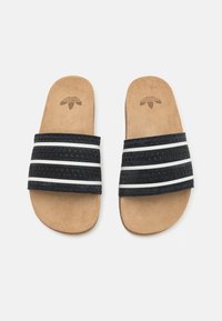 Svart plastikk-sandaler med tre teksturerte svarte stropper og hvite striper, med en lysbrun semsket skinn såle og en logo preget på innersålen.