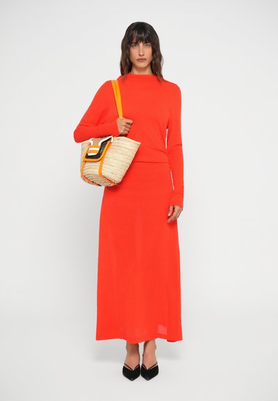 Abito lungo rosso a maniche lunghe con bodice aderente e gonna fluida. La modella porta una borsa in paglia intrecciata con dettagli arancioni e una chiusura nera a contrasto.