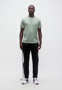 Άνδρας που στέκεται με ένα μπαστούνι γκολφ, φορώντας ένα ανοιχτό πράσινο μπλουζάκι, μαύρο παντελόνι και άσπρα παπούτσια γκολφ, μπροστά σε ένα απλό λευκό φόντο.