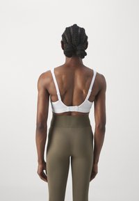 Hvit sports-bh med nettdetaljer, justerbare stropper og krokfeste, kombinert med høytaljede olivengrønne tights.