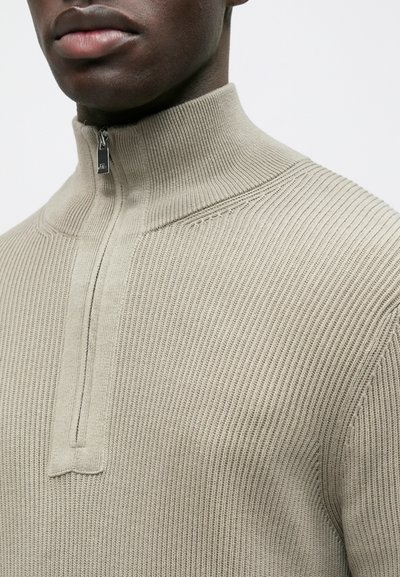 Pull beige en maille côtelée avec col montant et zip au quart. Présente des détails de couture subtils et un tissu texturé partout.