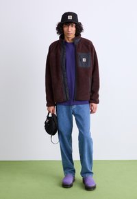 Brown fleece jacket with black accents, purple t-shirt, blue jeans, and purple clogs. Holding a small black bag. Wall is white, floor green.