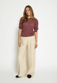 Nõelaga kootud burgundia punane kampsun tekstuurse rombikujulise mustriga, lühikeste varrukatega, koos kreemjate laia säärega pükstega ja tumepruunade jalanõudega.