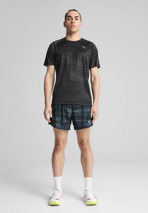 Maglietta sportiva nera a maniche corte con un sottile motivo a onde, abbinata a pantaloni corti blu scuro con strisce orizzontali più chiare. Scarpe da ginnastica bianche.