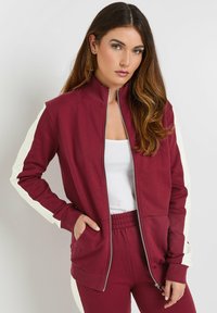 Giacca zip con collo alto di color marsala, pannelli laterali bianchi, tasche frontali e texture del tessuto morbida, abbinata a pantaloni marsala coordinati.