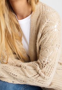 Femme blonde portant un cardigan beige en maille avec un motif de feuilles sur une chemise blanche, assise les bras croisés sur un jean bleu.