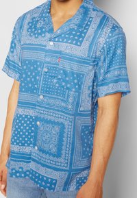 Niebieska koszula z krótkim rękawem w białe wzory paisley i geometryczne, z materiałowymi guzikami i kieszonką na piersi. Materiał wydaje się lekki i przewiewny.