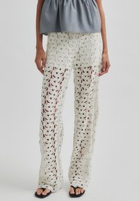 Pantaloni in pizzo all'uncinetto bianchi con un motivo floreale, caratterizzati da un design aperto e una vestibilità ampia. Abbinati a un top grigio e sandali neri.