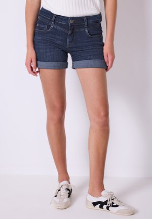 Denim shorts in dark blue with rolled cuffs, featuring front pockets, a button closure, and a slim fit design. Paired with white and black sneakers.
