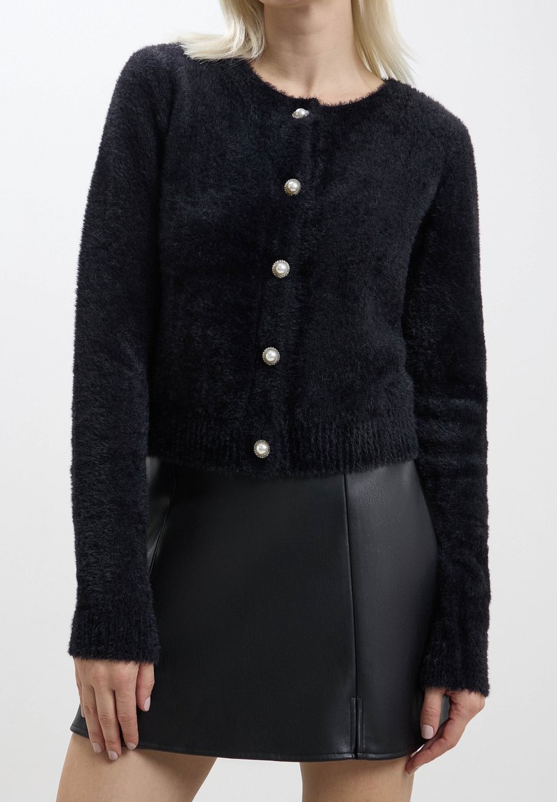 Femme portant un cardigan noir duveteux avec des boutons en perle et une mini-jupe en cuir noire, debout, les bras détendus le long du corps.