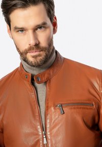 Giacca di pelle marrone con zip davanti, colletto a scatto e tasca sul petto con zip. Tessuto morbido con un design aderente e hardware minimale.