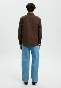 Braunes langärmliges Hemd mit Kragen, Schulterpasse und Knopfmanschetten, kombiniert mit hellblauen weit geschnittenen Jeans, vor einem schlichten Hintergrund.