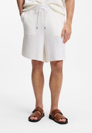 Mann trägt weiße, strukturierte Strickshorts mit Kordelzug und braune Ledersandalen, steht mit entspannten Händen.