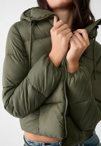Olivgrön, kort vadderad jacka med huva; har dragkedja, quiltad design och elastiska muddar för värme och komfort.