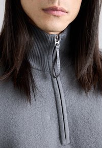 Pull en polaire grise avec un col côtelé haut et une demi-fermeture éclair. Le tissu semble doux avec une texture lisse. Tirette de fermeture éclair décorative visible.