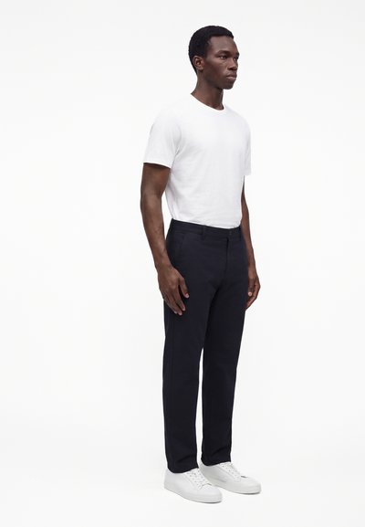 Homme debout portant un t-shirt blanc uni, un pantalon noir et des baskets blanches sur un fond blanc.