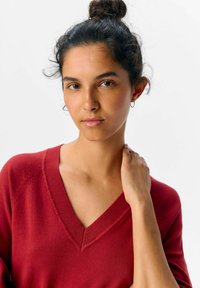 Czerwony sweter z dekoltem w serek, z żeberkowym wykończeniem, o miękkiej fakturze i rękawach raglanowych. Modelka ma upięte włosy w kok i delikatnie opiera rękę na ramieniu.