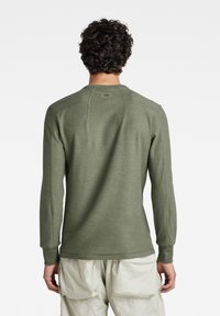 Långärmad skjorta i olivgrön tyg med en texturerad finish, som har rund halsringning och raglankragar, synlig sömnad och en baketikett.