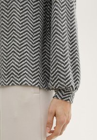 Suéter de punto con patrón de chevron en blanco y negro, con hombros caídos y puños elásticos, llevado sobre una tela de color beige claro.