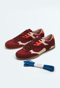 Paire de baskets bordeaux et beige avec des accents orange, des semelles marron, et un lot séparé de lacets bleus étiquetés "OYSHO" devant.