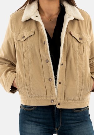Beige kordfløjlsjakke med hvid fleecekrave og for, båret over en sort top, kombineret med blå denimjeans.