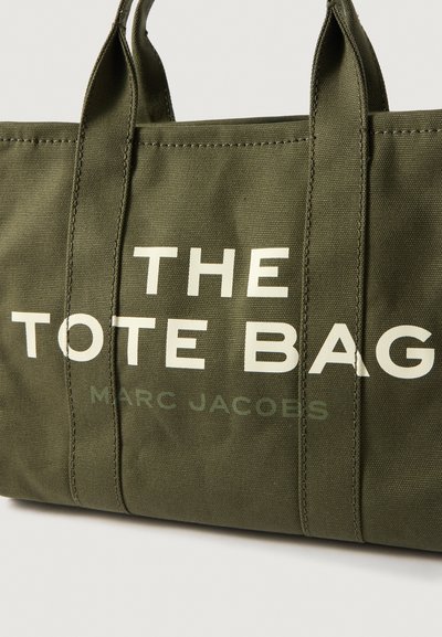 Geantă tote din pânză verde măsliniu cu text alb care citește "THE TOTE BAG" și "MARC JACOBS," având două mânere rezistente și cusături vizibile.