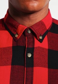 Červená a černá kostkovaná košile na knoflíky s špičatým límečkem, s mosaznými knoflíky a texturovanou látkou. Detailní pohled na výstřih.