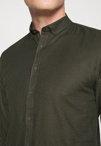 Camicia verde scuro con bottoni, realizzata in tessuto strutturato, con colletto appuntito, cuciture a contrasto e bottoni scuri.