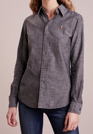 Femme portant une chemise grise ajustée boutonnée avec un petit logo brodé sur la poitrine et un jean bleu foncé, debout devant un fond uni.