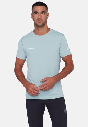 Mand iført lyseblå Mammut T-shirt og sorte bukser, stående med hænderne foldet foran kroppen, vendt mod kameraet på hvid baggrund.