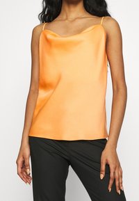 Orange satin cami med draperad halsringning och tunna spagettiband; slät textur och lös passform, i kombination med svarta byxor.