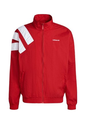 Crvena jakna sa patentnim zatvaračem napravljena od teksturiranog materijala. Ima bijele dijagonalne pruge na ramenu i izvezeni logo na grudima.
