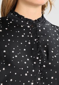 Blouse noire avec un motif d'étoiles blanches. Dotée d'un col à volants et d'une fermeture à boutons sur le devant. Texture satinée et lisse.