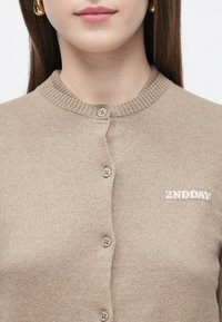 Beżowy dzianinowy kardigan z prążkowanym dekoltem, z pięcioma guzikami i białym haftem "2ND DAY" po lewej stronie. Miękka w dotyku.