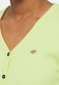 Ljusgrön ribbad cardigan med V-ringning, svarta knappar och en liten färgglad logotyp på vänster sida.