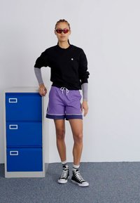 Schwarzer Pullover, lila Shorts mit marineblauer Umrandung, schwarze High-Top-Sneaker und rote Sonnenbrille. Steht neben einem blauen Aktenschrank.