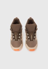 Ζευγάρι καφέ και μπεζ παπούτσια Adidas Terrex για πεζοπορία με πορτοκαλί σόλες, επάνω μέρος από δίχτυ και κορδόνια, σε απλό ανοιχτό φόντο.