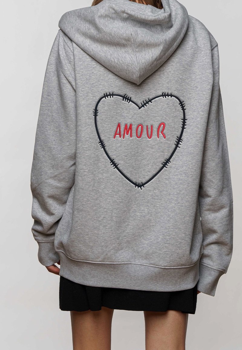 Sweat à capuche gris avec un grand contour de cœur brodé et "AMOUR" en rouge, avec des détails cousus en noir. Texture douce, coupe décontractée.