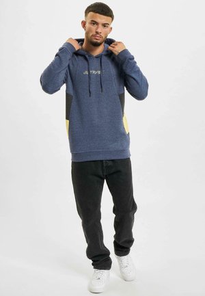 Navy blue hoodie with contrasting black side panels and yellow accents, featuring a drawstring hood and embroidered text. Paired with dark jeans.