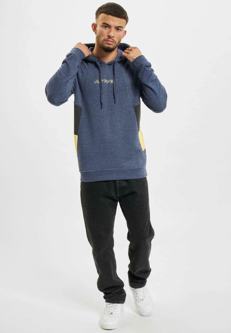 Sudadera de color azul marino con paneles laterales en negro contrastante y acentos amarillos, con capucha ajustable y texto bordado. Combinada con jeans oscuros.