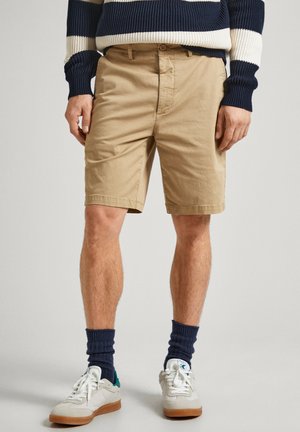 Pantalones cortos de algodón beige con corte recto, bolsillos frontales y trabillas para cinturón. Combinados con zapatillas blancas y calcetines de canalé azul marino.