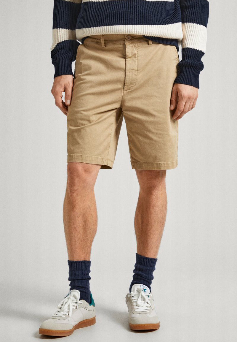 Pantaloni corti in cotone beige dal taglio dritto, con tasche frontali e passanti per cintura. Abbinati a sneaker bianche e calze a coste blu navy.