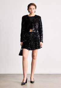 Svart paljettblazer och minikjolset med en kort silhuett, i kombination med svarta högklackade skor och en liten handväska.