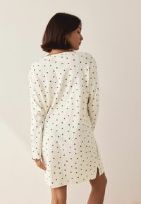 Valkoinen neulemekko, jossa on punainen sydänkuvio, ribbikuviointi, pitkät hihat ja sivuvetoketjut. Pyöreä kaula-aukko ja aaltoilevat hihansuut.