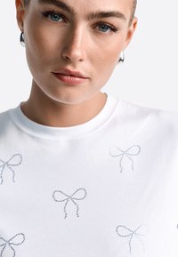 Biała bawełniana koszulka z wieloma niebieskimi haftowanymi wzorami w kokardy. Posiada klasyczny okrągły dekolt i krótkie rękawy. Gładka faktura.