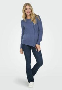 Kék kötött pulóver laza szabással, bordás mandzsettákkal és aljjal; sötét denim harangszárú farmerral és fehér sportcipővel párosítva. Egyszerű dizájn.
