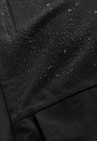 Tejido negro resistente al agua con una textura suave, presentando gotas de agua visibles. El diseño incluye costuras sutiles y un acabado mate.