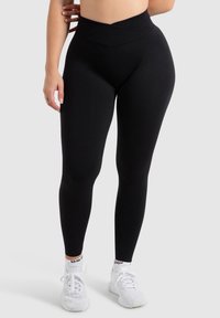 Zwarte high-waisted leggings met een gladde textuur en een crossover taillebandontwerp, gecombineerd met witte sportschoenen.